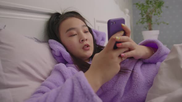 Happy Millennial Asian Woman Typing Message Using Mobile Phone Lying in Bed alt