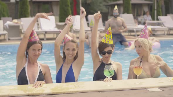 Positive Relaxed Caucasian Women Dancing in Water at Poolside with Blurred Multiracial Men Sitting alt