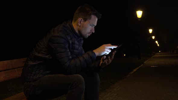 A Young Handsome Man Sits on a Bench and Works on a Smartphone in an Urban Area at Night alt