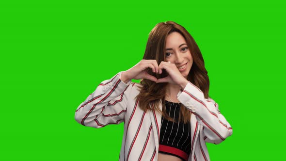 Adorable young woman posing on green screen showing heart and thumb up gestures alt