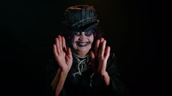 Frightening Senior Woman with Halloween Witch Makeup Waves Hand Palm in Hello Hi Gesture Welcomes alt