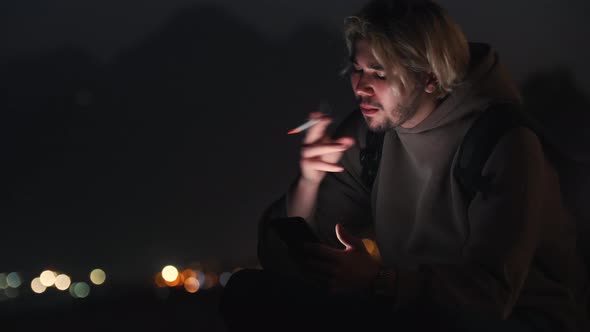 Blonde Man in Beige Hoodie Sitting on the Night Beach and Smoking a Cigarette alt