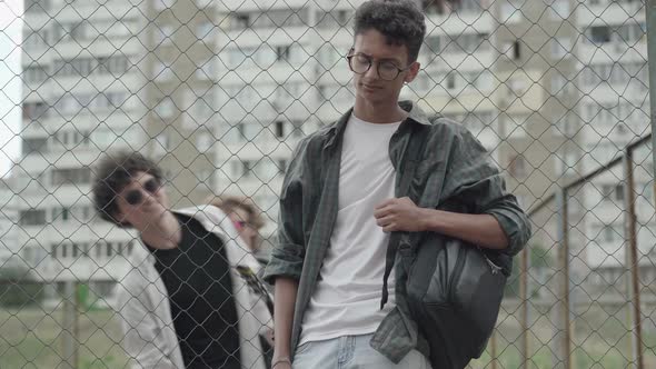 Portrait of Young African American Man Standing at Mesh Fence As Caucasian Boys Walking at the alt