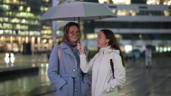 Two Women are Standing Under an Umbrella Laughing and Chatting Against the Background of a Blurred alt