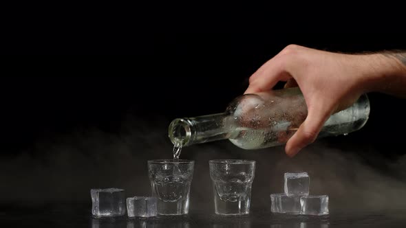 Hand Pours Vodka Tequila or Sake From Bottle Into Shot Glasses on Black Background with Ice Cubes alt
