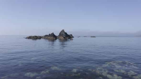 Low aerial skims water surface toward rugged eroded sea rocks in ocean alt