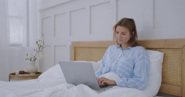 Young Millennial Girl Sitting on a Bed in the Morning, Uses Laptop Computer. Caucasian Woman Home alt