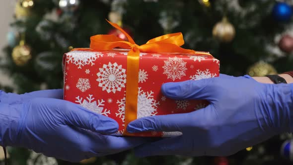 Giving a Gift Box in Protective Gloves with Christmas Tree on a Background.