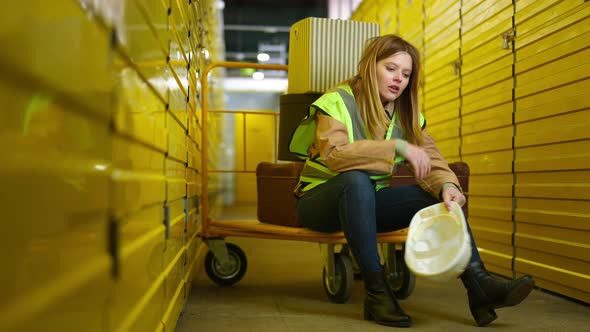 Wide Shot Exhausted Young Woman Sitting on Industrial Trolley Taking Off Hard Hat alt