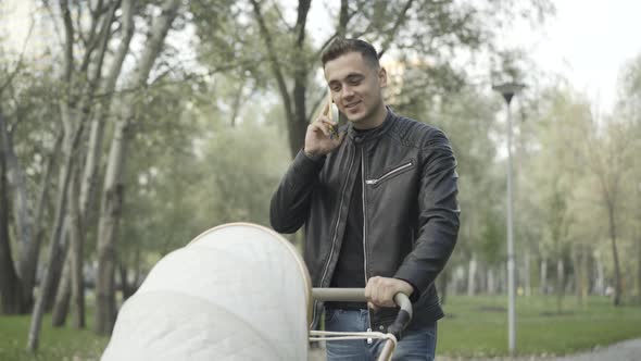 Happy Smiling Young Caucasian Father Walking with Baby Stroller in Park and Talking on the Phone alt