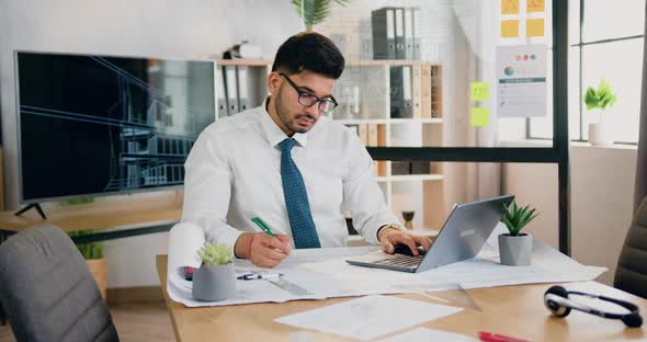 Bearded Worker in Glasses writing Notes on Blueprint Comparing Datas with Information on Laptop alt