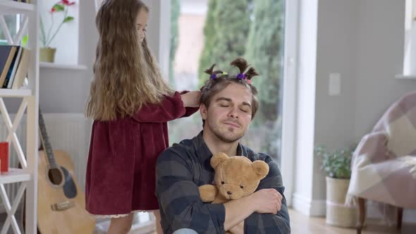 Concentrated Little Girl Making Ponytails on Hair of Joyful Father and Pulling Parent Ears alt