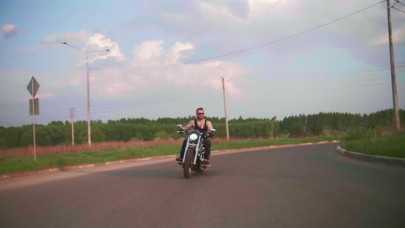 Stylish Biker with Tattoos Rides a Motorcycle on a Country Road at Sunset