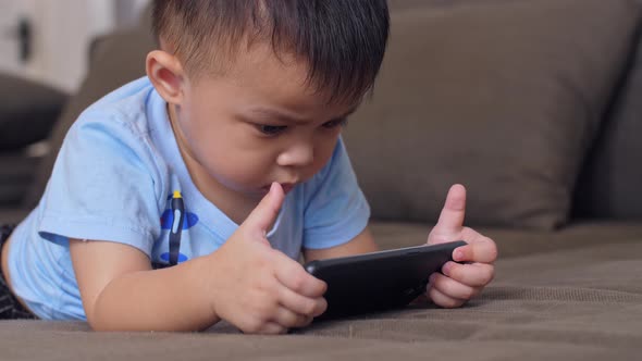 Asian Little Boy Laying On A Sofa Plays With Smartphone alt