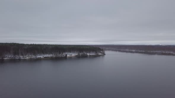 Aerial footage of russian lake in winter. Drone goes up above the lake. Cloudy weather. Snowy trees.