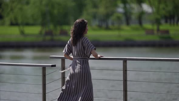 the Brunette in a Striped Dress Stands By the River on a Sunny Day alt