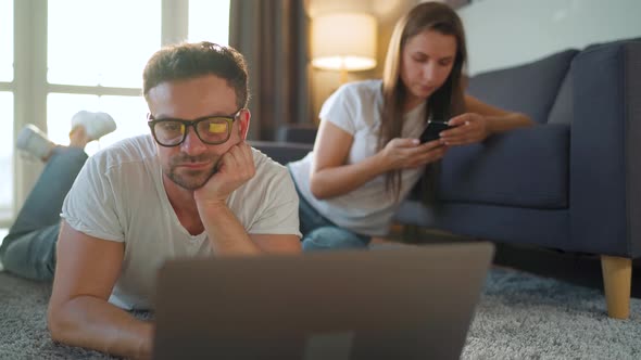 Couple Spends Time Together in a Cozy Room: Man Lies on a Carpet and Uses a Laptop, Woman Using alt