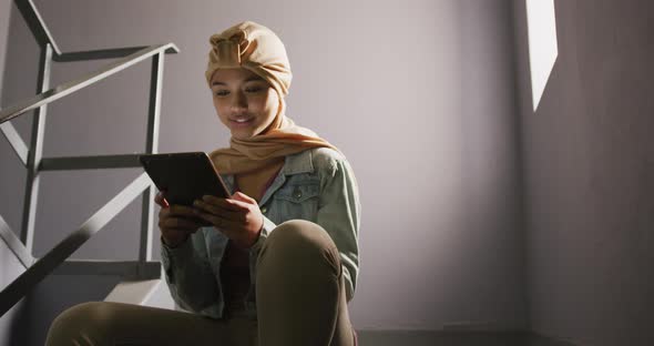 Asian female student wearing a beige hijab sitting on stairs and using a tablet alt