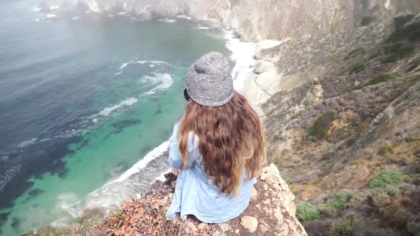 A young woman sitting on top of a cliff with the Bixby Creek Bridge in the background. alt