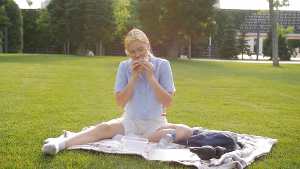 A Teenage Girl in the Park on the Grass Eating Sandwiches Washes It Down with Water From a Plastic alt