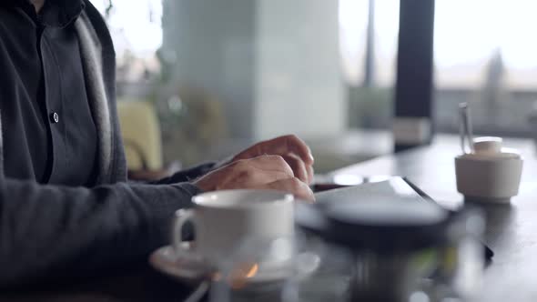 Close-up Shot of a Guy Chatting Online on a Tablet in Cafe, Cup of Tea on Table alt