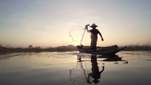 Silhouette fisherman throwing fishing net during sunset with boats at the lake.