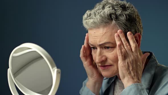 An Elderly Grayhaired Woman Carefully Examines Her Reflection in the Mirror Lightly Touching the