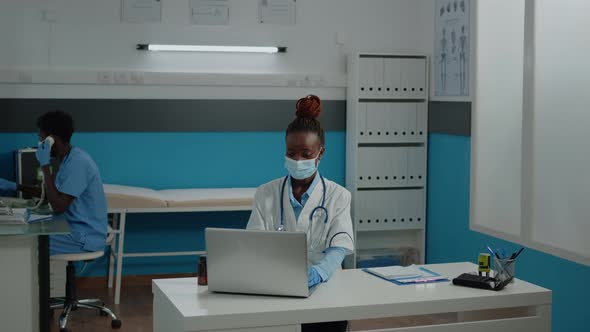 Portrait of African American Doctor Sitting at Desk with Face Mask alt