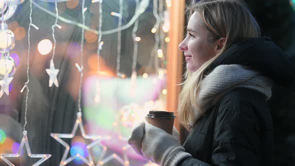 Young happy girl with a cup of hot coffee looks into the glowing window of the store.