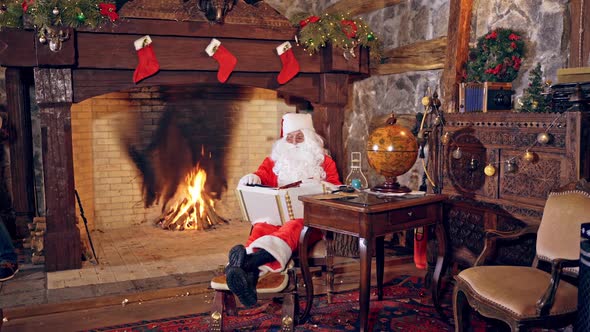Santa Claus is sitting in specially decorated room and looking at the book. alt