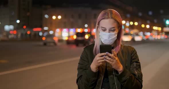 Woman Wearing Mask Looks in Smartphone Against Car Lights alt