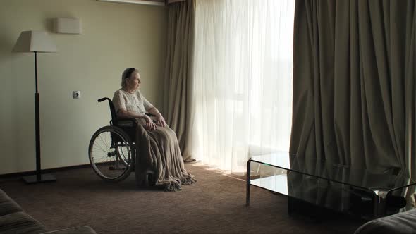 Senior Woman Sit on Wheelchair and Looks Through the Window alt
