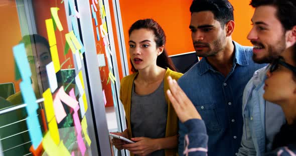 Smiling executives discussing over sticky notes on glass wall alt