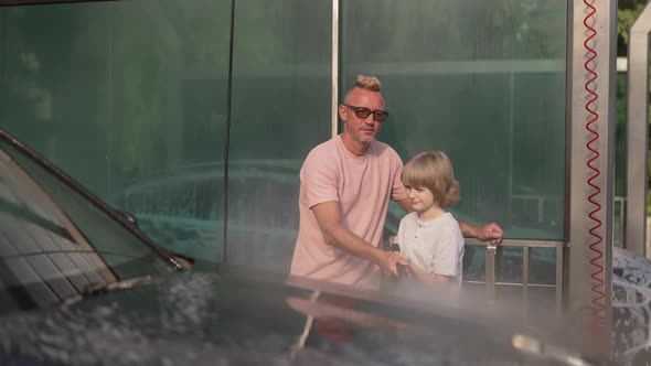 Front View Portrait of Positive Caucasian Father and Son Cleaning Vehicle Spraying Water at Camera alt
