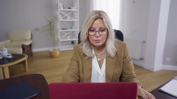 Front View Portrait Focused Concentrated Woman in Eyeglasses Sitting at Table with Laptop Reading alt