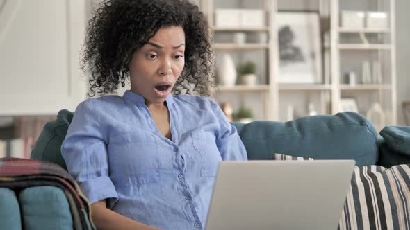 Shocked African Woman Using Laptop Sitting on Couch alt