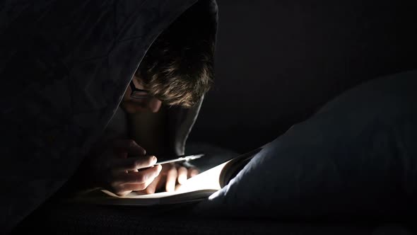 Man Reads a Book with a Flashlight Under the Covers at Night