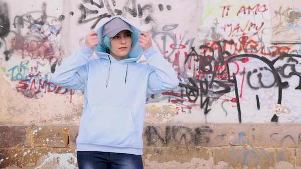 Blue haired teenage girl wearing light blue hoodie and stays against graffiti wall alt