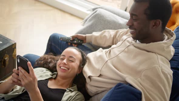 Overhead Shot Of Relaxed Young Couple At Home On Sofa Watching TV And Checking Mobile Phone