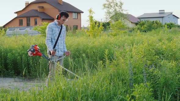 Man Mows Weeds Around the House with a Trimmer alt