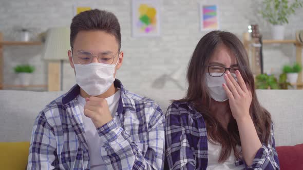 Sick of a Cold Young Sian Couple Sitting in Protective Medical Masks on the Couch alt