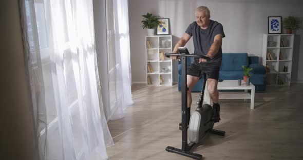 Grey Haired Man Is Training on Spinning Bike in Living Room Healthy Lifestyle at Middle Age alt