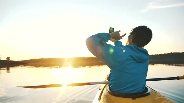 Man Makes Selfie on Smartphone While Sitting in Kayak on Calm Lake at Sunset, Outdoor Activities alt