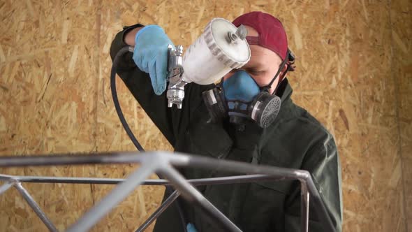 Worker in a Red Cap and a Green Jacket Paints Metal Products With Airless Spray. Slowmotion