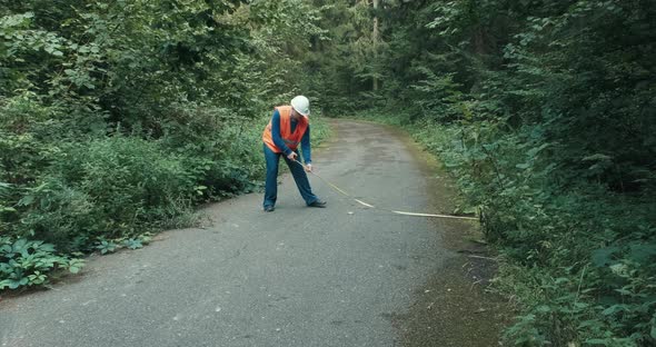 Worker in Workwear and on Highway in Forest Measures Roadway with Tape Measure