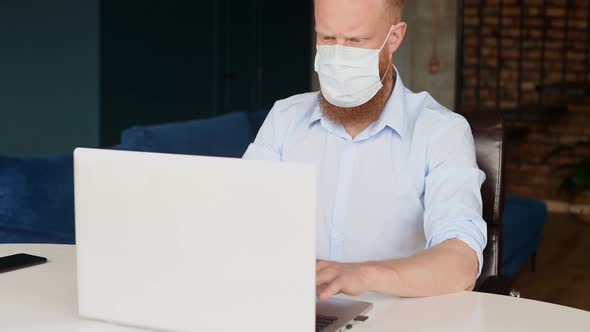 Concentrated Redhead Bearded Man Wearing Protective Medical Mask Using Laptop