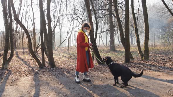 Pedestrian Walkway Woman in Orange Coat and Yellow Hoodie with Face Mask Walk with Dog in the Park alt