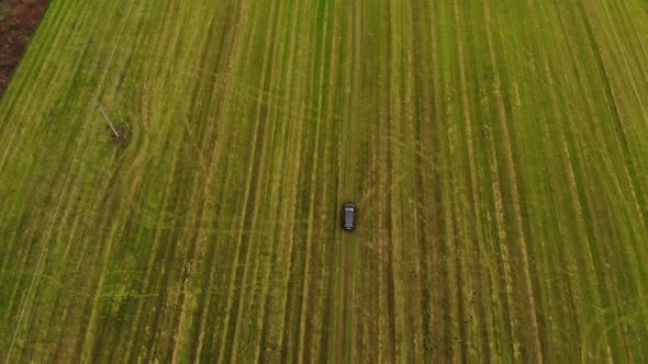 Back Aerial View at Black Car Moves Along Ground Road Crossing Green Fields