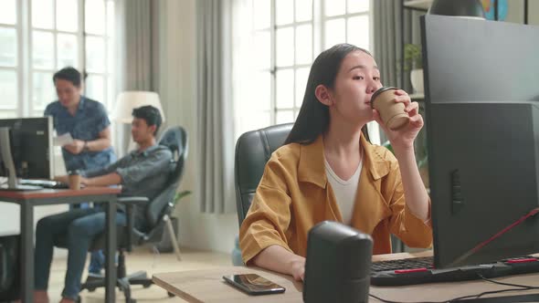 Asian Female Drinking Coffee While Works On Her Personal Computer With Big Display In Office alt
