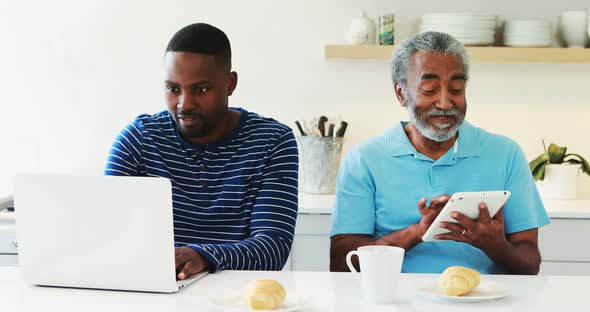 Father and son using laptop and digital tablet in kitchen alt
